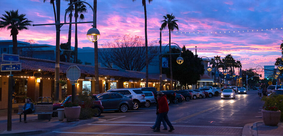 street view at sunset of lit single story shops with people crossing the road and cars parked and moving along the road.