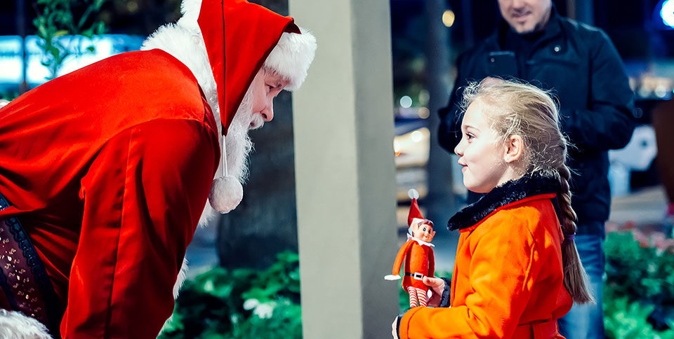 Outdoor scene showing Santa leaning down to talk to an animated young girl with braided blond hair, an orange coat holding an elf on the shelf while a smiling man in a black jacket is photographing the scene