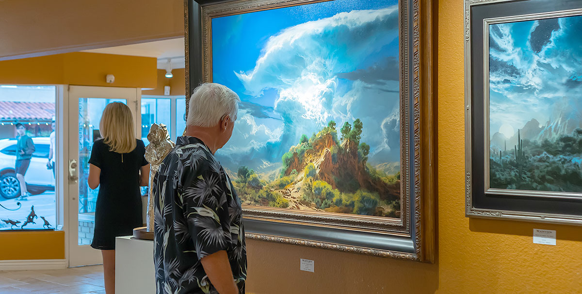 Man in a dark patterned shirt and white short hair looking at a large dramatic landscape painting in a gallery with dark mustard colored walls