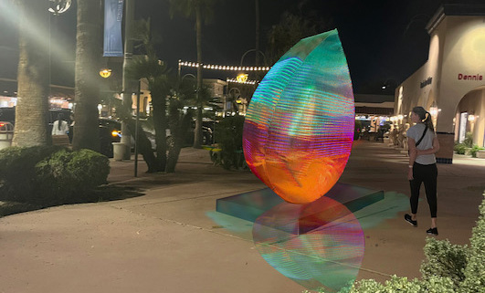 8 foot tall multi-colored teardrop shaped internally light sculpture on pavement sidewalk with a woman walking by at night