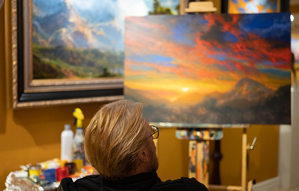 Blond haired man sitting in front of a western landscape painting in progress with brilliant sunset sky