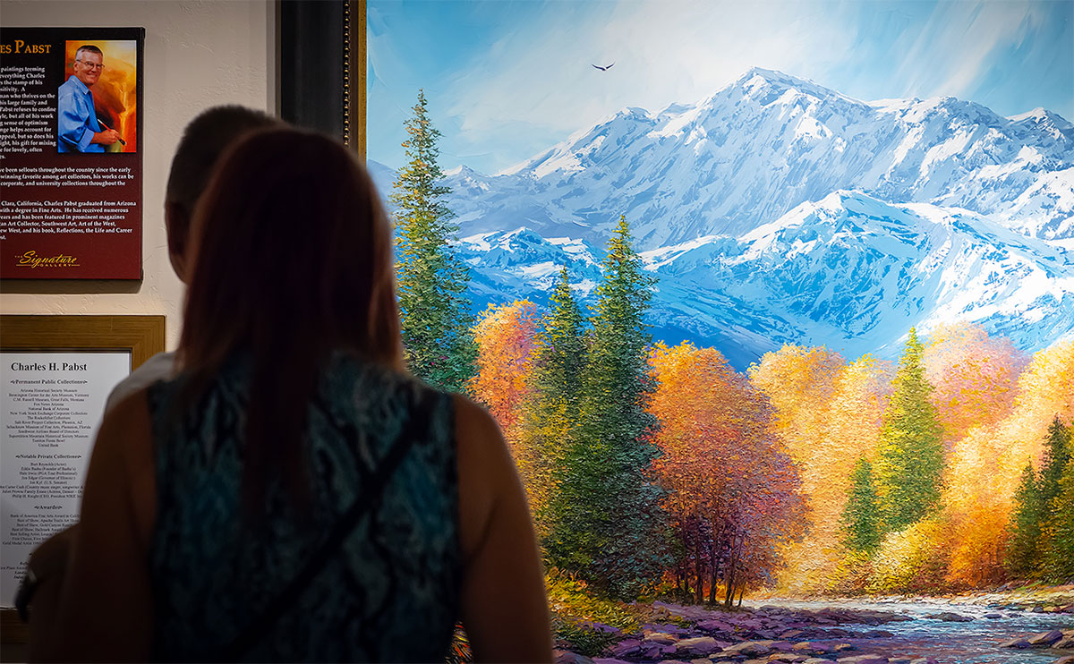 Woman looking at a painting of trees in fall foliage along a rocky stream with rugged snowy mountains in background