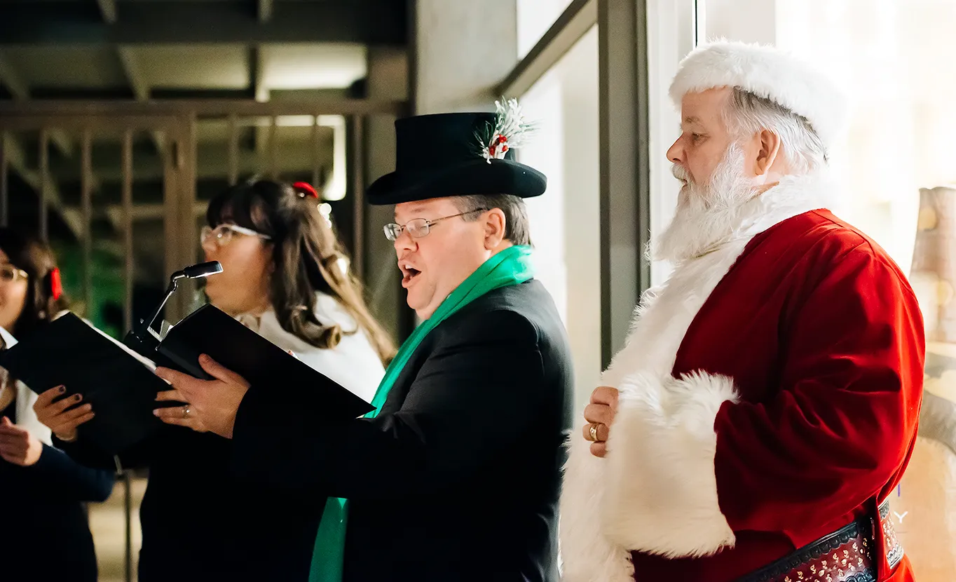 Santa Claus singing with carolers dressed in black and green.