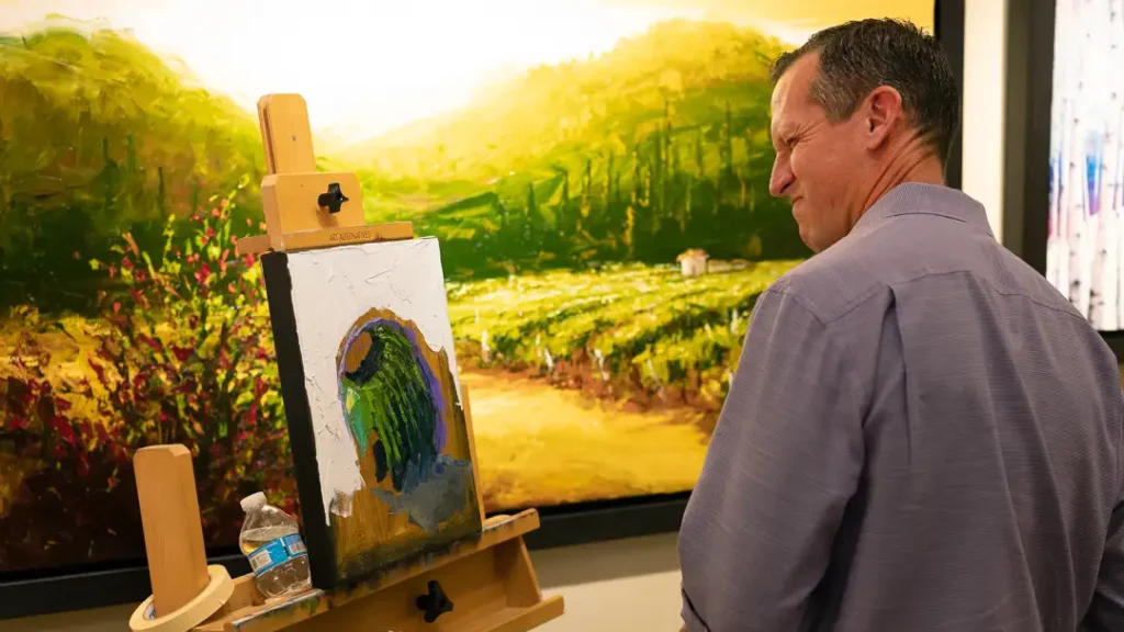 White man in a collared button down shirt grimacing at a painting of a cactus on an easel with finished landscape paintings on a wall in the background.