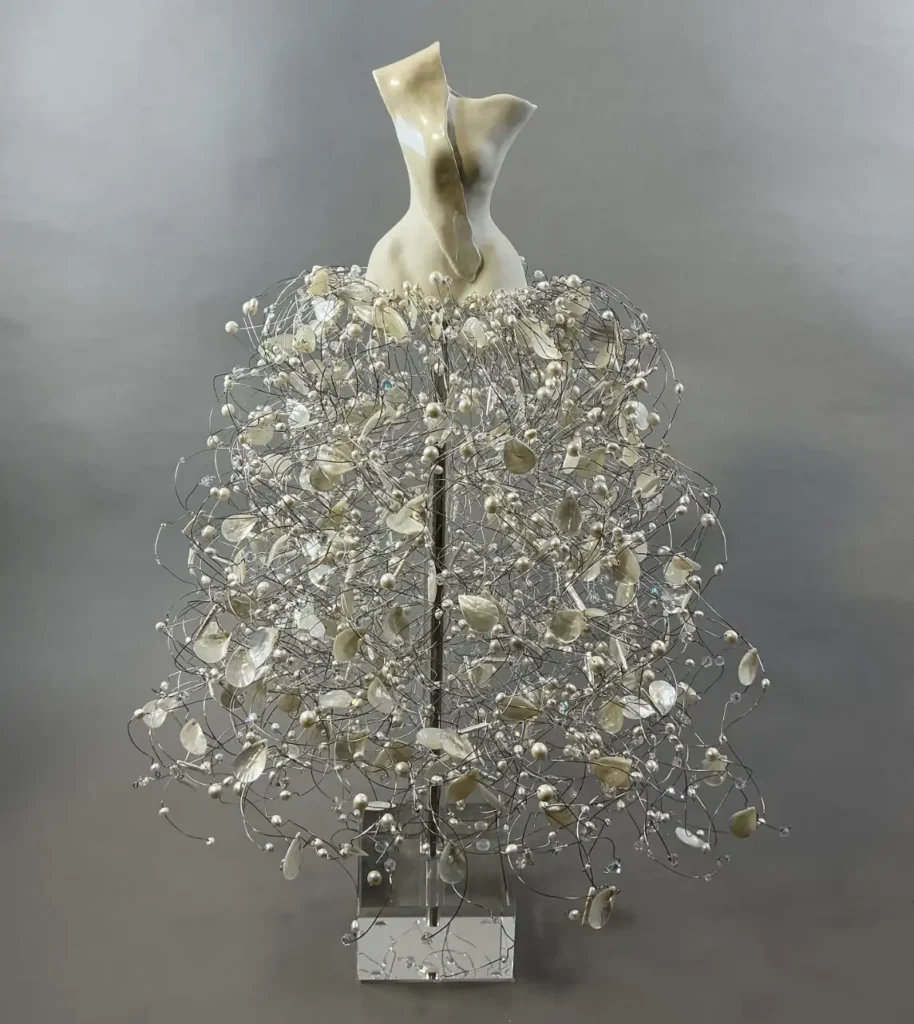 Sculpture by Estella Fransbergen showing a porcelain cream colored abstract female torso/bodice with a large flowing skirt-like arrangement of silver wire and cream colored shells and pearls.
