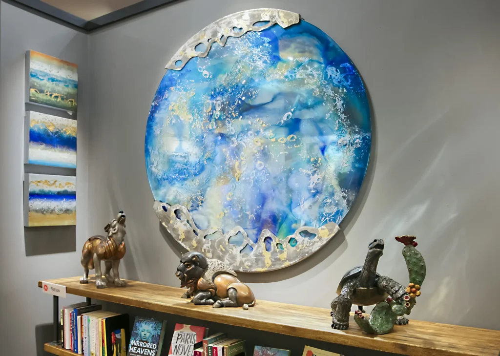 Interior wall decorated with a large round ocean-like color scheme invoking brilliant turquoise water, wave foam and golden flecks of sunlight framed with formed and pierced metal edging above a low wooden bookcase with sculptures.